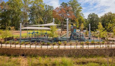 A playground with large grey shade sails overhead surrounded by landscaping and a black fence.