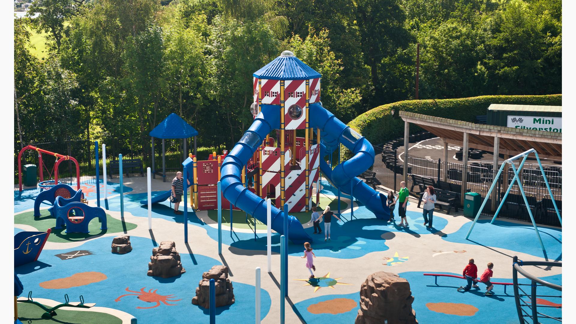 Elevated view of a nautical themed playground with a large two leveled tower designed with striped red and white siding panels and two blue tunnel slides.