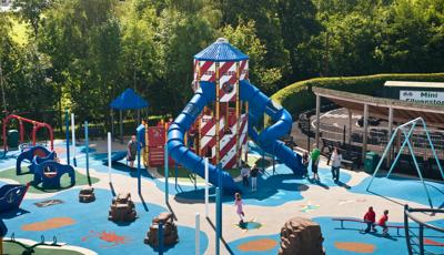 Elevated view of a nautical themed playground with a large two leveled tower designed with striped red and white siding panels and two blue tunnel slides.