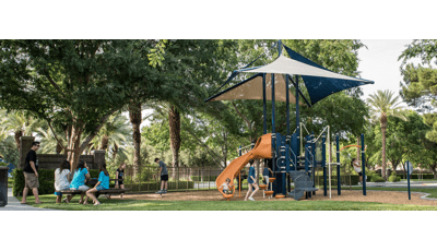 A little boy in an orange and white stripe shirt sits at the bottom of the slide as a girl stands in front of the the play structure. Adults sit on benches near by.