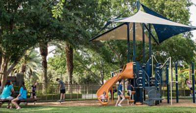 A little boy in an orange and white stripe shirt sits at the bottom of the slide as a girl stands in front of the the play structure. Adults sit on benches near by.