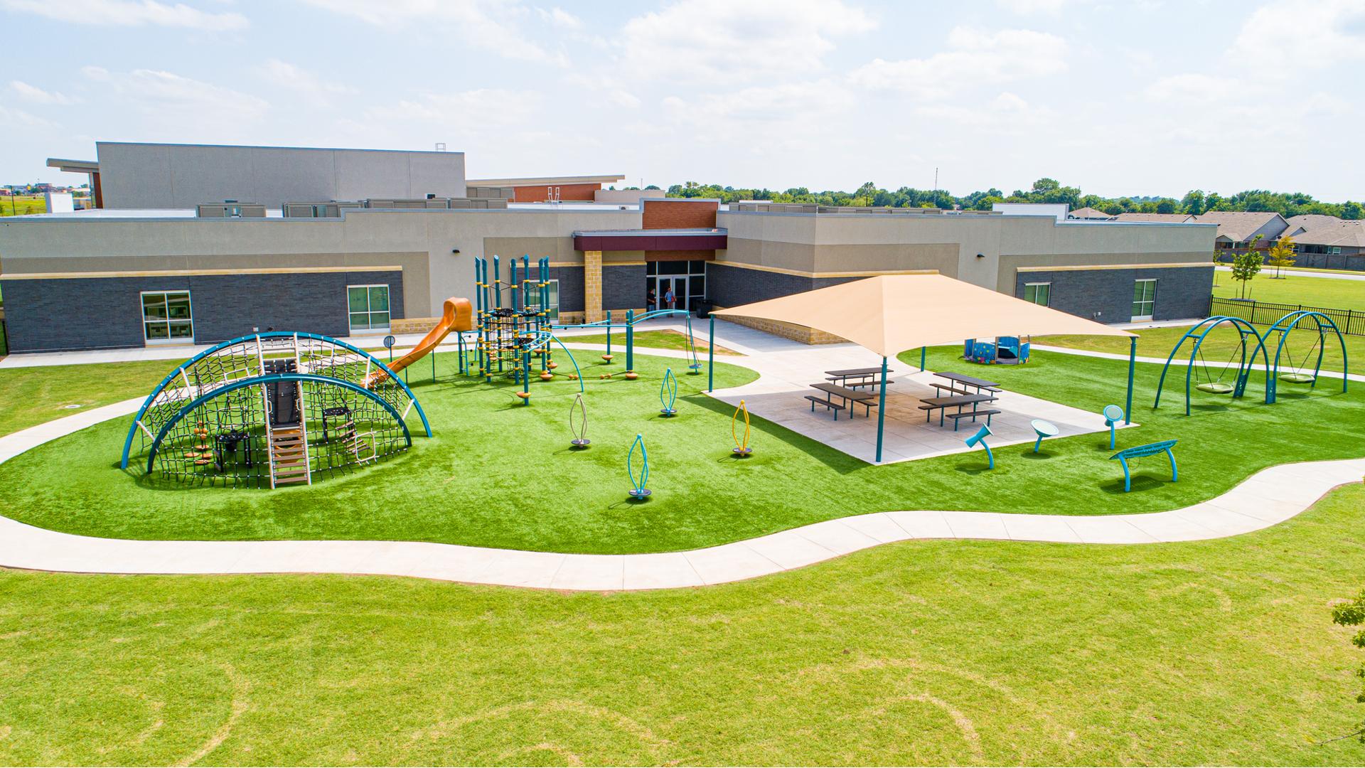 Elevated view of an outdoor school playground with play structures standing spinners, swing set, and outdoor musical instruments. A small meeting area with picnic tables sits in the middle of the play area under a large tan square shade.