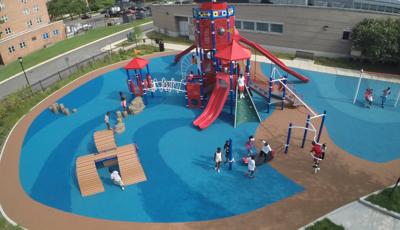 Aerial view of a red light-house themed playground with children playing. Surfacing mimics blue water and brown sand with buildings in the background. 