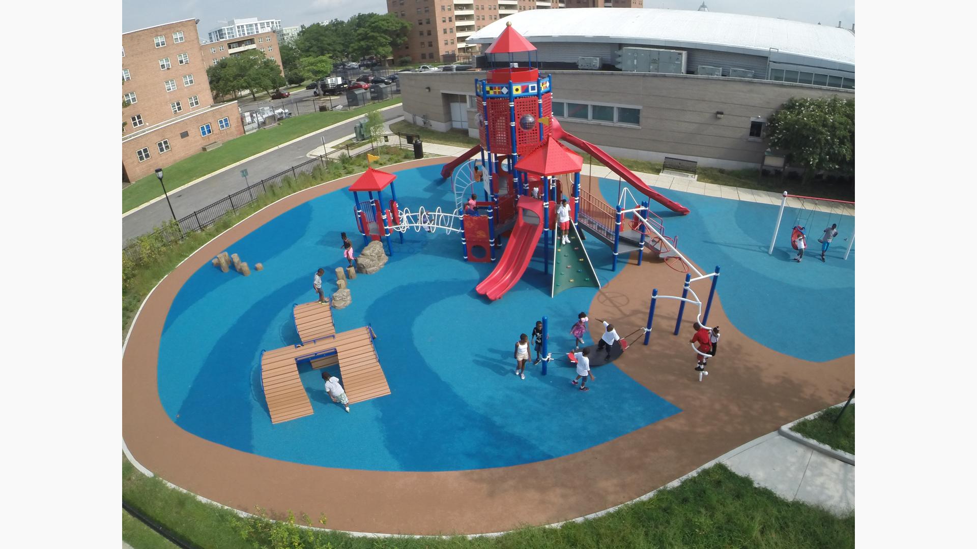 Aerial view of a red light-house themed playground with children playing. Surfacing mimics blue water and brown sand with buildings in the background. 