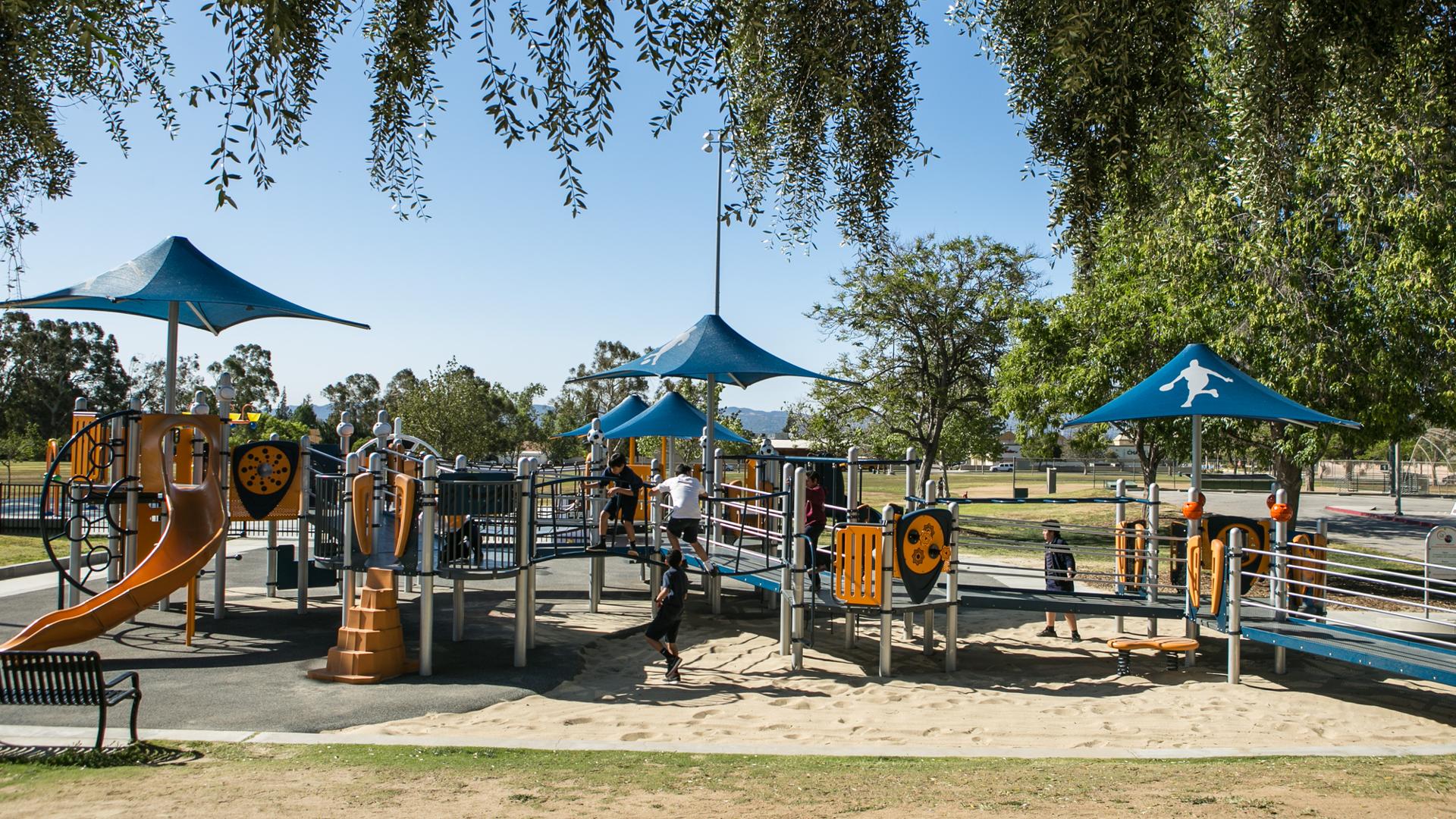 Children playing on sports themed playground.  Blue shade with a custom sports figure cover part of the playground.  The playground includes ramps so kids of all abilities can reach the play elements. 