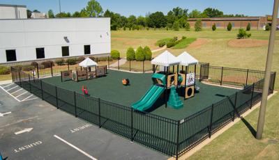 A fenced in play area has two play structures for toddler aged children set on the side of a building.