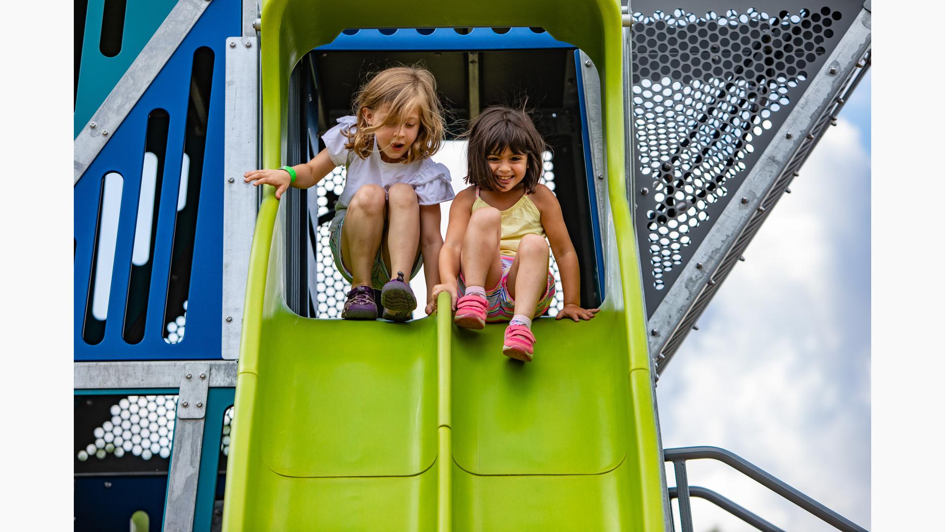 A Little Girl Sliding Down A Green Slide At The Playground. by