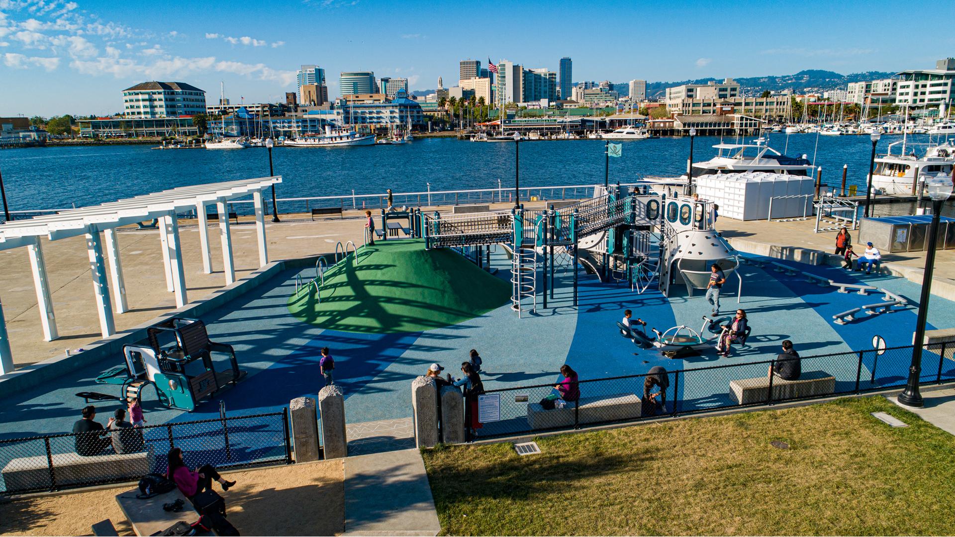 Families play on a custom boat themed play structure with additional surrounding play activates set next to a waterway with the Oakland skyline in the background.