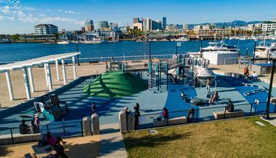 Families play on a custom boat themed play structure with additional surrounding play activates set next to a waterway with the Oakland skyline in the background.