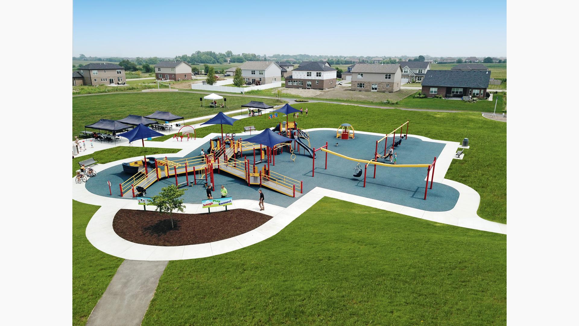 Elevated view of a neighborhood park inclusive play area. The playground is connected by accessible ramps and bridges surrounded by a swing set, zip line, and manual carousel. 