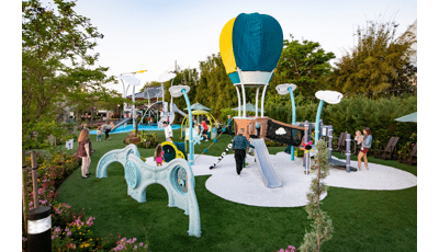 Families play on a hot air balloon themed play structure in the foreground as others play on a larger air themed play structure in the background. The play area is surrounded by lush gardens and trees.