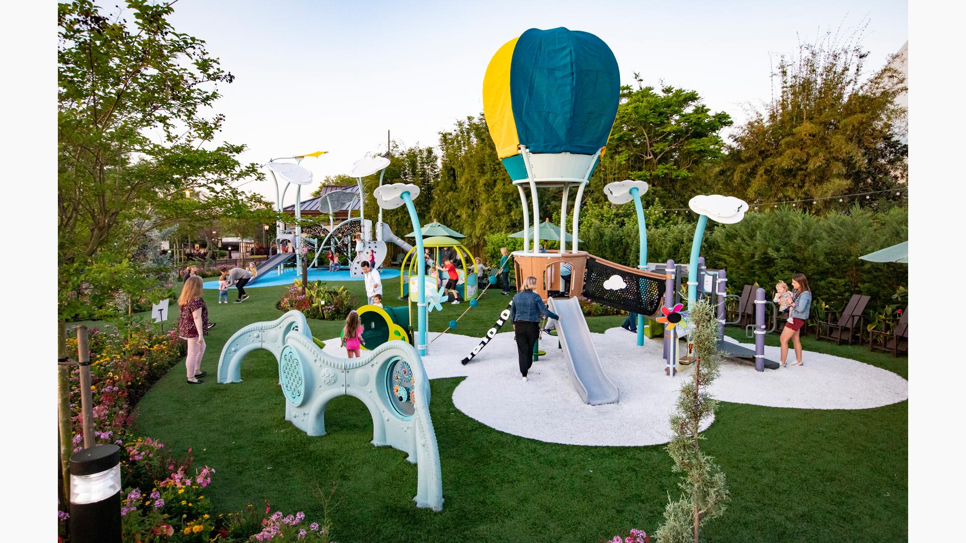 Families play on a hot air balloon themed play structure in the foreground as others play on a larger air themed play structure in the background. The play area is surrounded by lush gardens and trees.