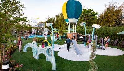 Families play on a hot air balloon themed play structure in the foreground as others play on a larger air themed play structure in the background. The play area is surrounded by lush gardens and trees.