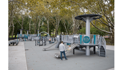 A mother stands by watching as her daughter slides down a metal dual lane slide on a futuristic designed inclusive playground surrounded by a grove of trees. 