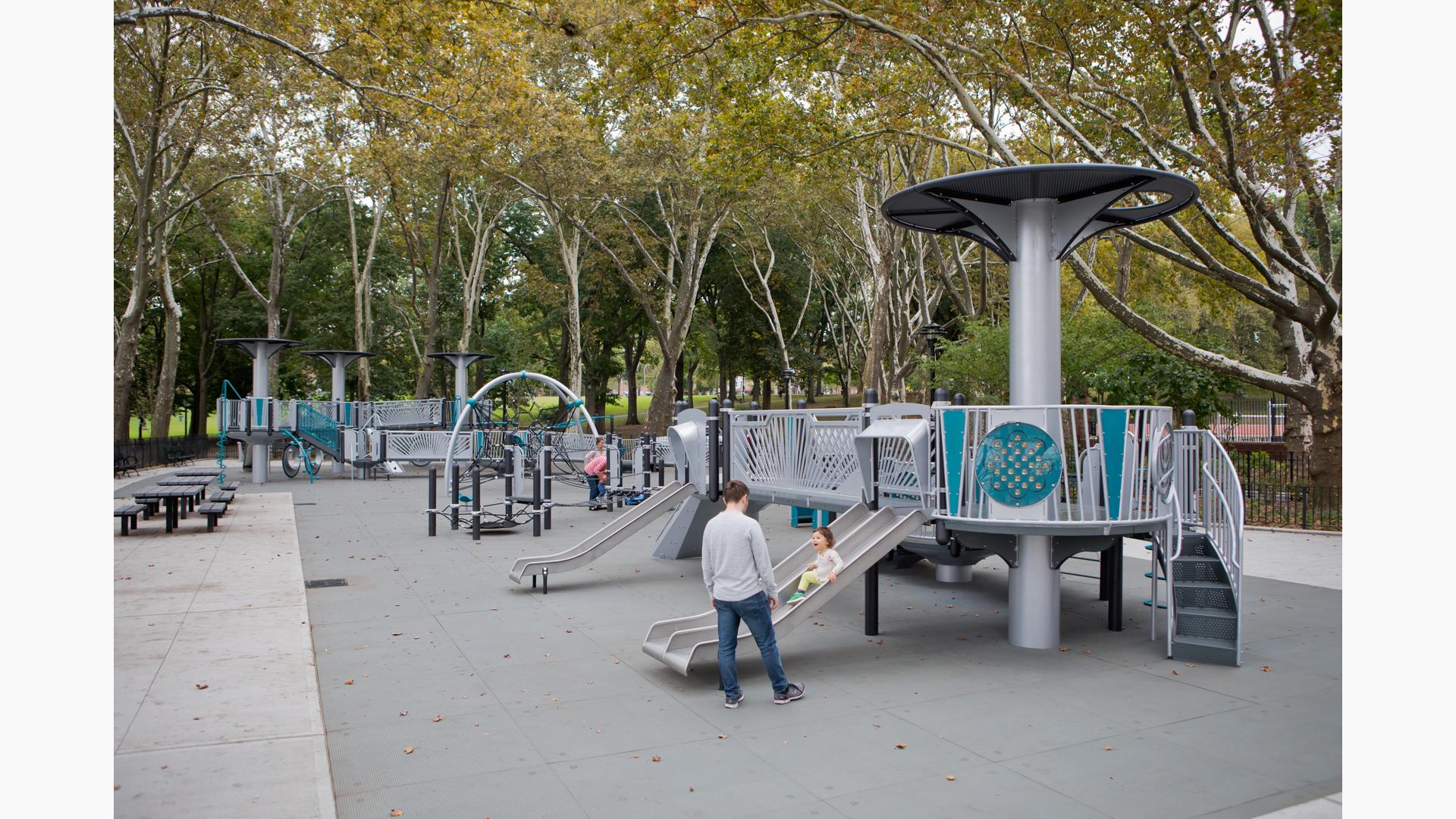 A mother stands by watching as her daughter slides down a metal dual lane slide on a futuristic designed inclusive playground surrounded by a grove of trees. 