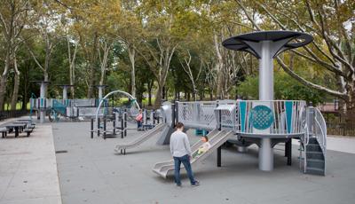 A mother stands by watching as her daughter slides down a metal dual lane slide on a futuristic designed inclusive playground surrounded by a grove of trees. 