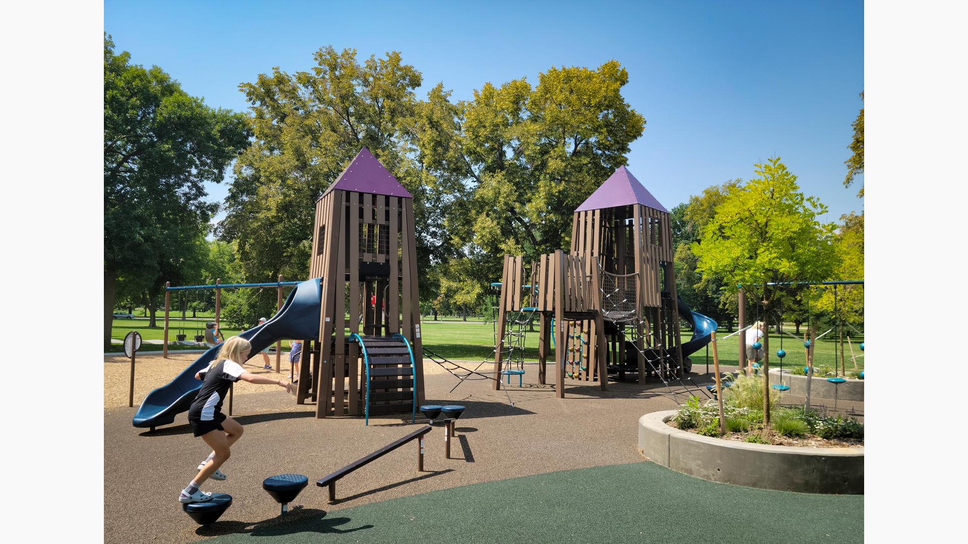Two play structures connected by a rope ladder climber are designed like castle towers made up of recycled plastic lumber at an outdoor park.