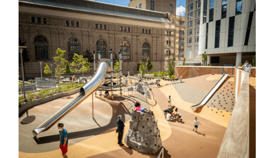 People enjoying the sun in Waterline Square. A little girl in a pink shirt sits on top of a rock climbing structure, while a boy climbs a net on a Netplex.