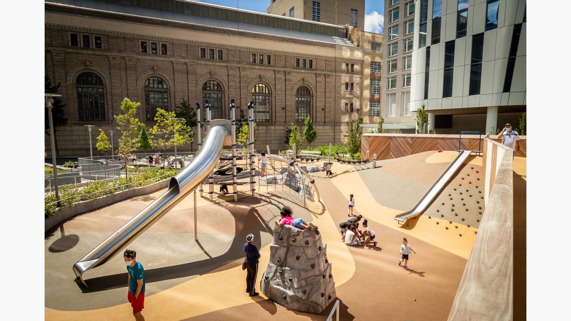 People enjoying the sun in Waterline Square. A little girl in a pink shirt sits on top of a rock climbing structure, while a boy climbs a net on a Netplex.