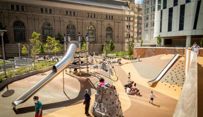 People enjoying the sun in Waterline Square. A little girl in a pink shirt sits on top of a rock climbing structure, while a boy climbs a net on a Netplex.