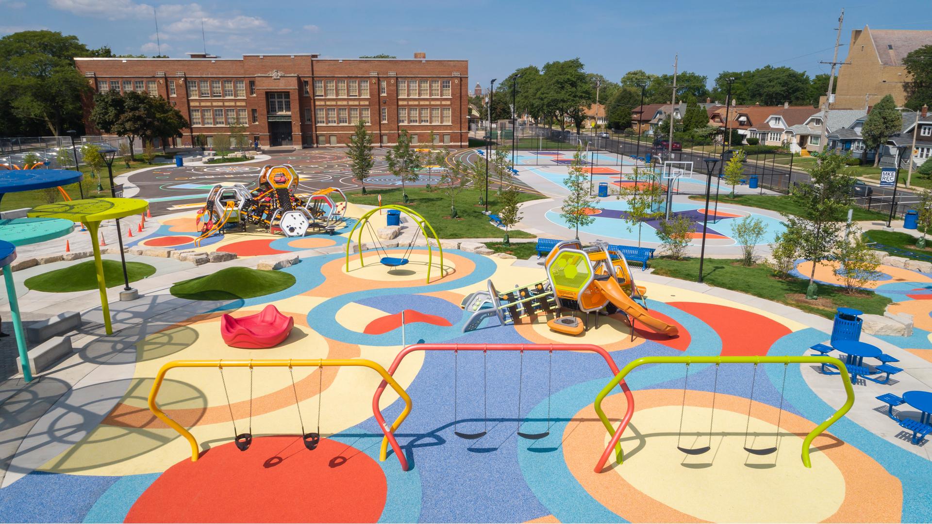 Elevated view of a large play area with colorful safety surfacing, brightly colored swing sets and two separate play structures made up of hexagonal shaped play pods. Basketball courts sit beyond the playground area with a large brick building in the background. 