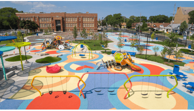 Elevated view of a large play area with colorful safety surfacing, brightly colored swing sets and two separate play structures made up of hexagonal shaped play pods. Basketball courts sit beyond the playground area with a large brick building in the background. 