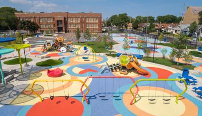 Elevated view of a large play area with colorful safety surfacing, brightly colored swing sets and two separate play structures made up of hexagonal shaped play pods. Basketball courts sit beyond the playground area with a large brick building in the background. 