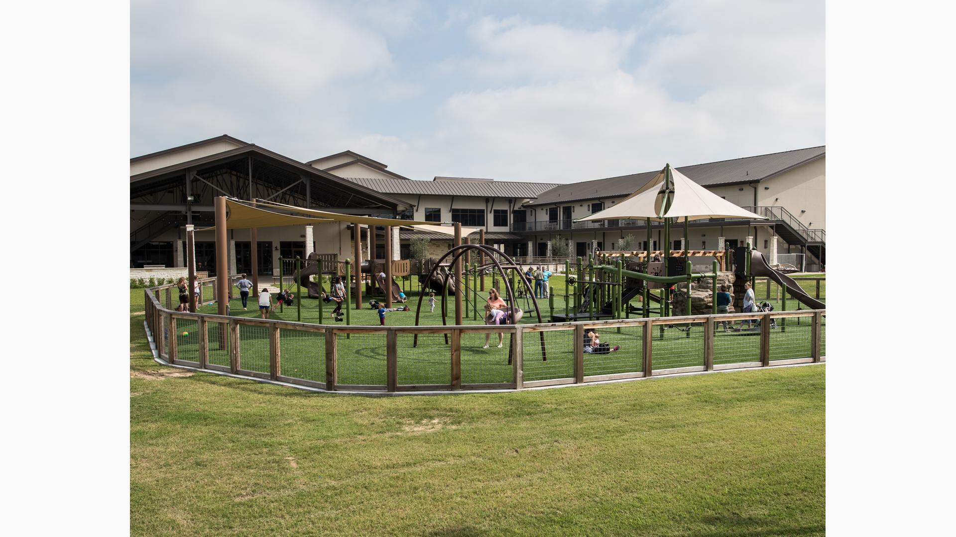 Families play on a sunny day in green grass at Grace Bible Church. SkyWays shade canopies covering parts of the playground. The church looms in the background.