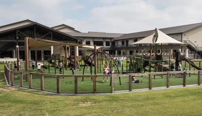Families play on a sunny day in green grass at Grace Bible Church. SkyWays shade canopies covering parts of the playground. The church looms in the background.