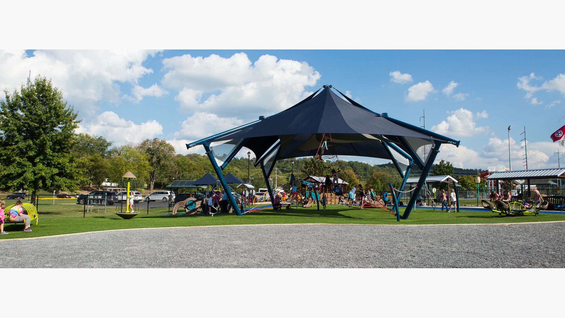 A crowd of children climb on a pyramid rope climber underneath a large navy blue peaked shade as other children play on surrounding see saws, spinners, and play panels.  