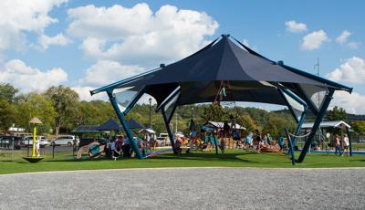 A crowd of children climb on a pyramid rope climber underneath a large navy blue peaked shade as other children play on surrounding see saws, spinners, and play panels.  