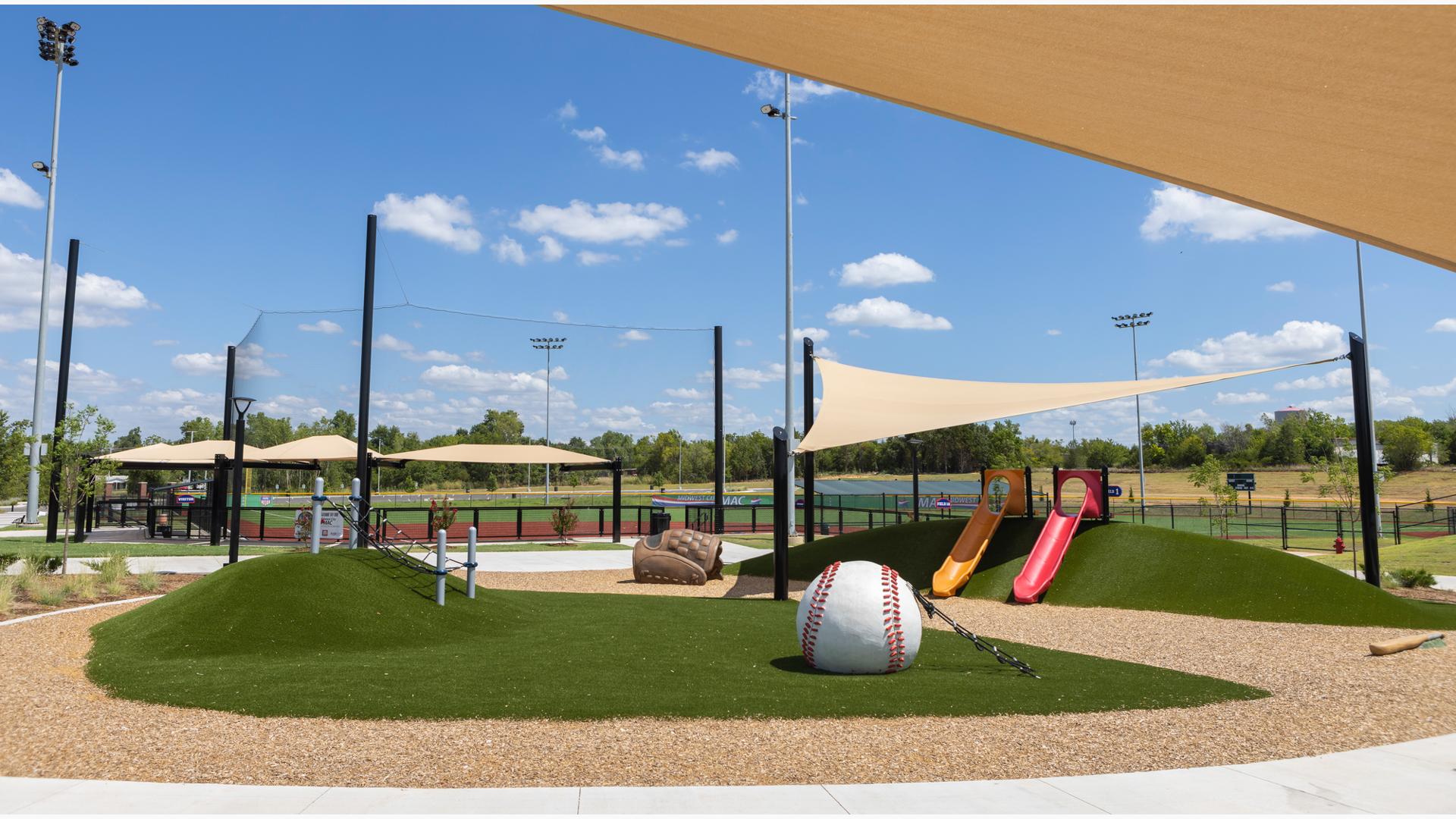 A small play area with green artificial grass sits amongst baseball fields with rope climbers, slides, and a custom designed baseball glove crawl tunnel and baseball climber. 