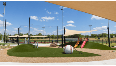 A small play area with green artificial grass sits amongst baseball fields with rope climbers, slides, and a custom designed baseball glove crawl tunnel and baseball climber. 