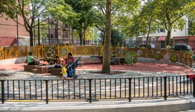 A group o fpeople sit on wooden benches under tree canopies, as a little girl in a yellow shirt runs by on the playground. On the play structure the boys stand at the top of the stairs together.