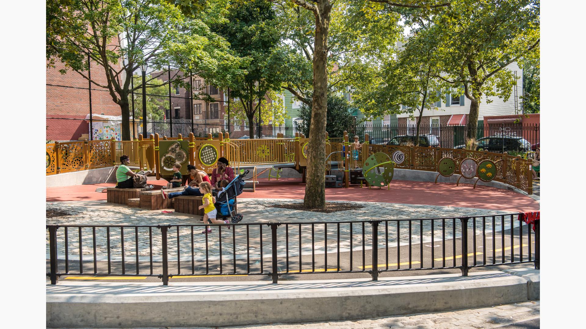 A group o fpeople sit on wooden benches under tree canopies, as a little girl in a yellow shirt runs by on the playground. On the play structure the boys stand at the top of the stairs together.