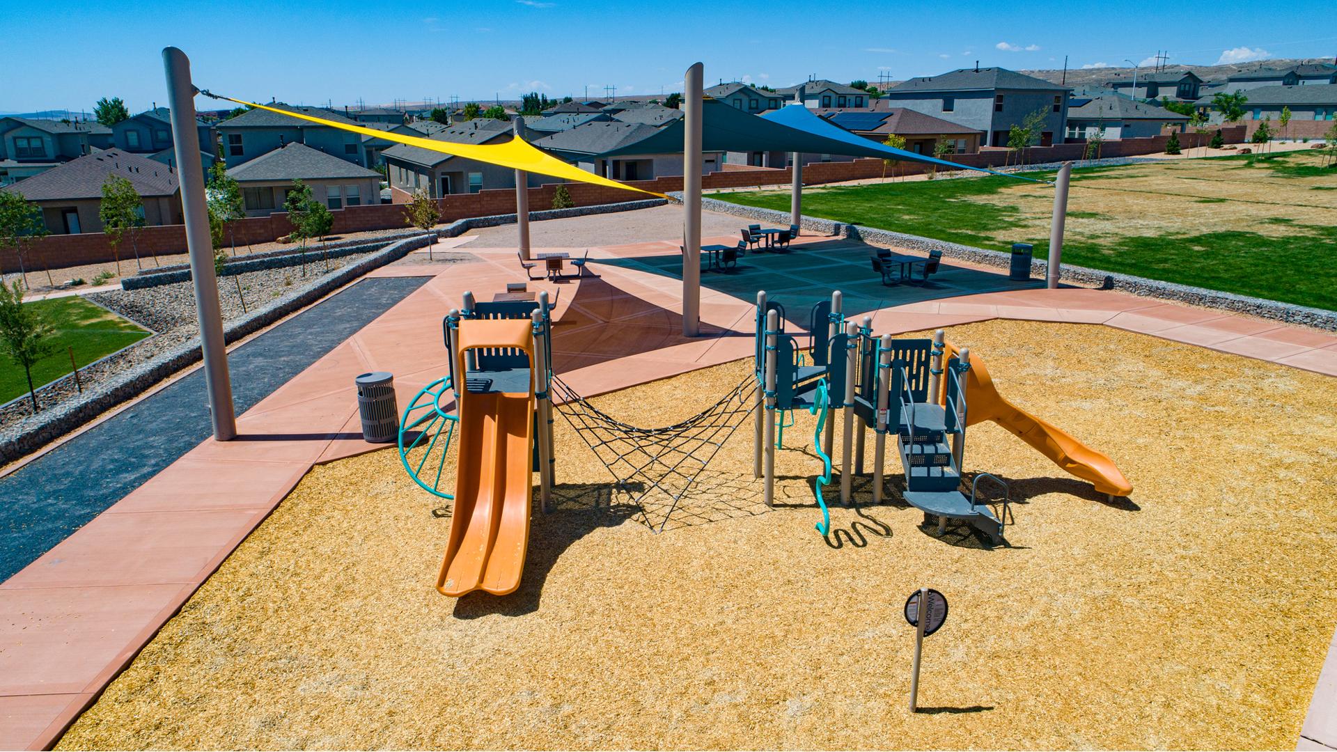 Elevated view of a playground with two large triangular shades in the background cover a sitting picnic area with tables.