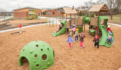 Kids play on  PlayBooster play structure at Early Childhood Family Center. It's a cloudy day. A boy in an organge jacket walks on the log balance beam. The PlayShaper play structure is in the background.