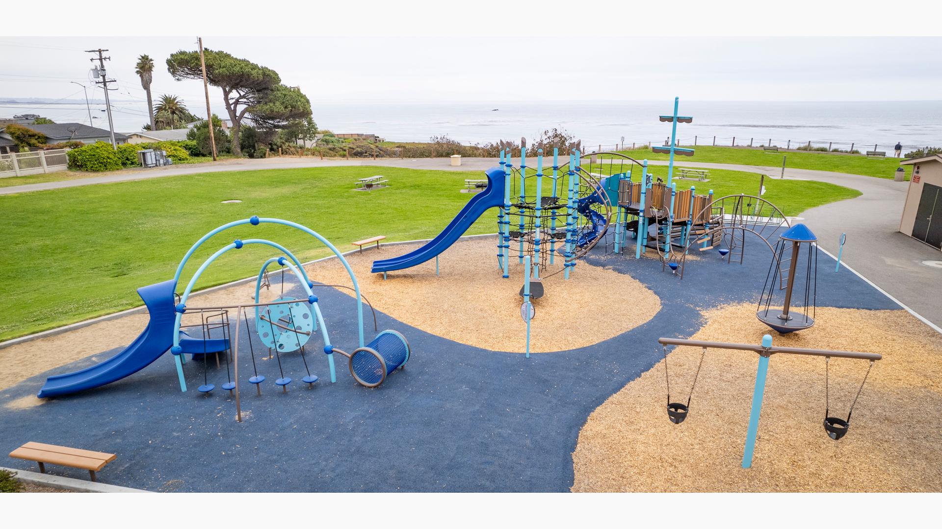 A play area with a large play structure for older children styled like an imaginative ship with a secondary play area for younger children with toddler swings nearby. A wide-open grassy play area sits behind the playground as the entire park sits on the top of a bluff overlooking the ocean.