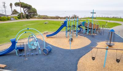 A play area with a large play structure for older children styled like an imaginative ship with a secondary play area for younger children with toddler swings nearby. A wide-open grassy play area sits behind the playground as the entire park sits on the top of a bluff overlooking the ocean.