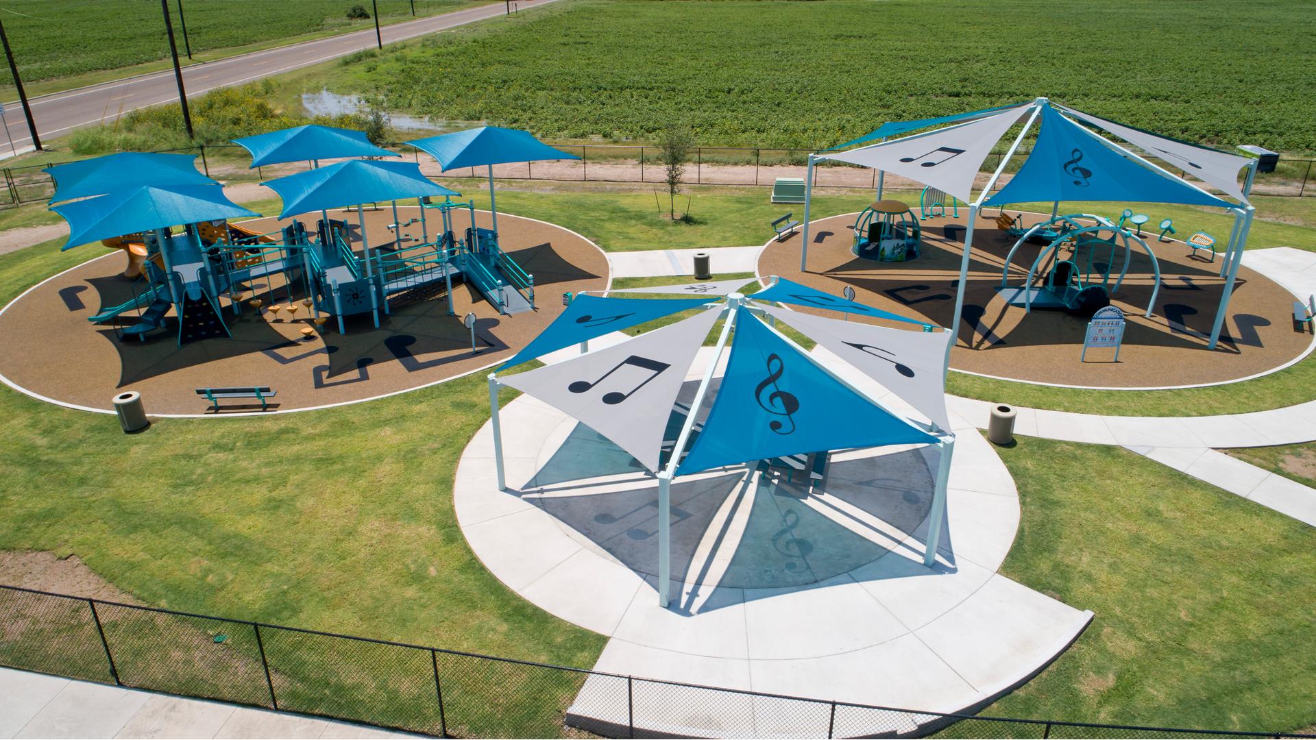 Elevated view of a large fenced in play area with three separate play locations. Two of the play locations have a large hexagonal shape shade system with large music notes and treble clefs printed on the light grey and blue shade panels. The third play area has five square blue shades protecting the large inclusive play structure below from the sun. 