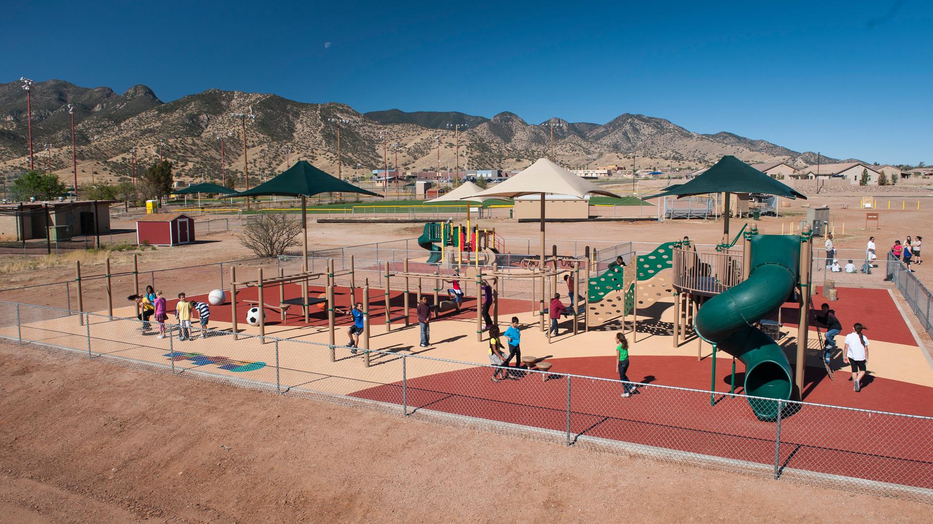 With a mountain range in the background, children play at Sierra Vista Children's Fitness Park. Climbing the PlayBooster play structure. Each area is covered by CoolToppers shade systems.