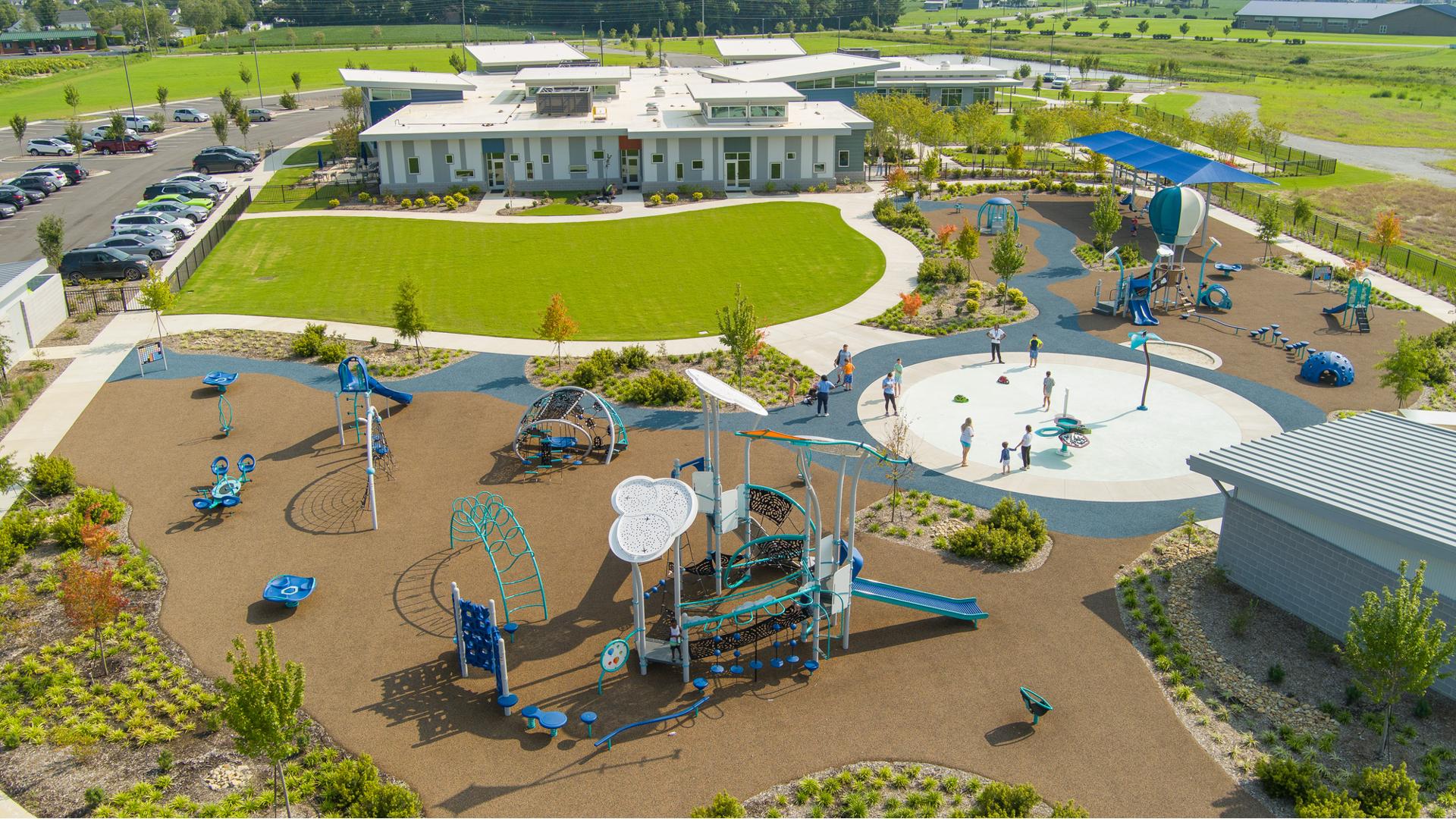 Elevated view of a inclusive playground and splash pad next to a building and grassed open gathering area.