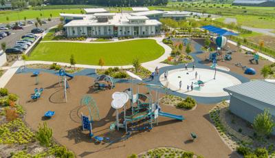 Elevated view of a inclusive playground and splash pad next to a building and grassed open gathering area.