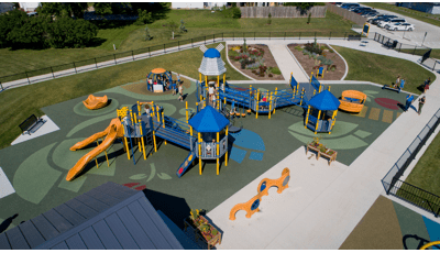 Elevated view of an inclusive Dutch themed playground surrounded by a black iron fence. The safety surfacing is designed with tulip shapes, a roof top like a windmill, and children cut outs dressed in Dutch costumes. Children play on a manual carousel next to the large play structure.