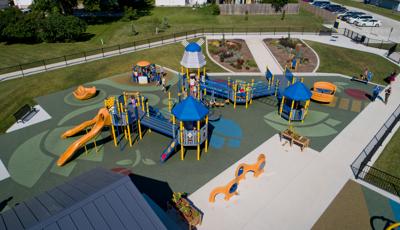 Elevated view of an inclusive Dutch themed playground surrounded by a black iron fence. The safety surfacing is designed with tulip shapes, a roof top like a windmill, and children cut outs dressed in Dutch costumes. Children play on a manual carousel next to the large play structure.