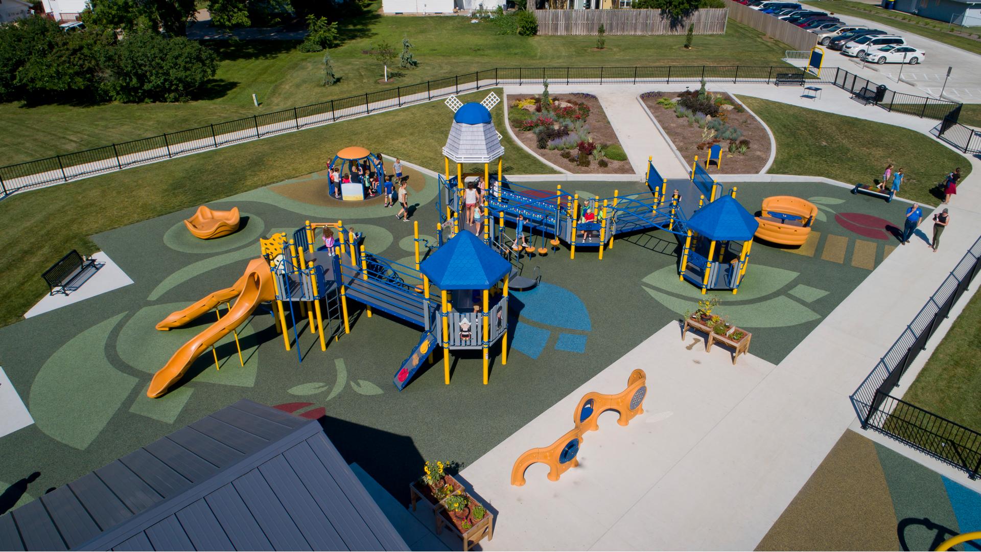 Elevated view of an inclusive Dutch themed playground surrounded by a black iron fence. The safety surfacing is designed with tulip shapes, a roof top like a windmill, and children cut outs dressed in Dutch costumes. Children play on a manual carousel next to the large play structure.