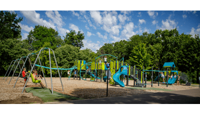 Children play all over a large playground with multiple climbers, slides and play panels. A long line of swing sets sit to he left of the play structure as lush green trees surround the play area. 