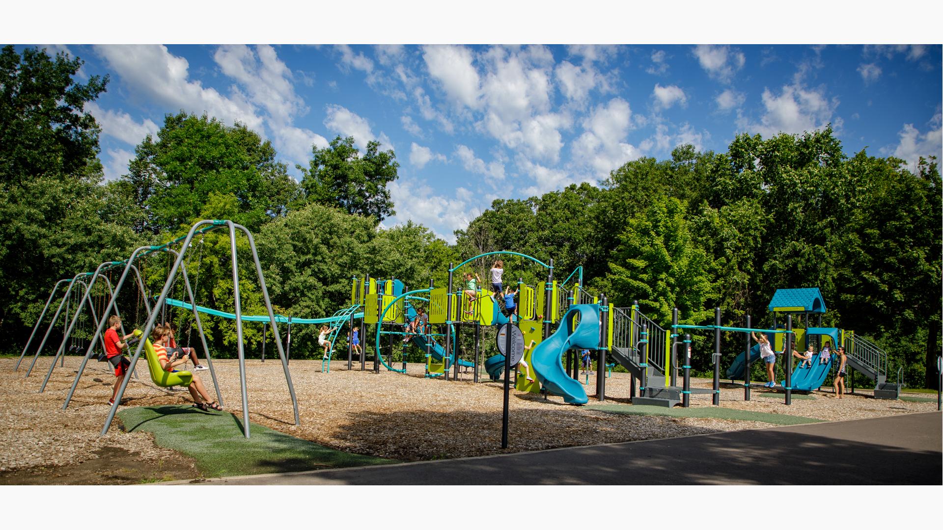 Children play all over a large playground with multiple climbers, slides and play panels. A long line of swing sets sit to he left of the play structure as lush green trees surround the play area. 