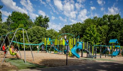 Children play all over a large playground with multiple climbers, slides and play panels. A long line of swing sets sit to he left of the play structure as lush green trees surround the play area. 
