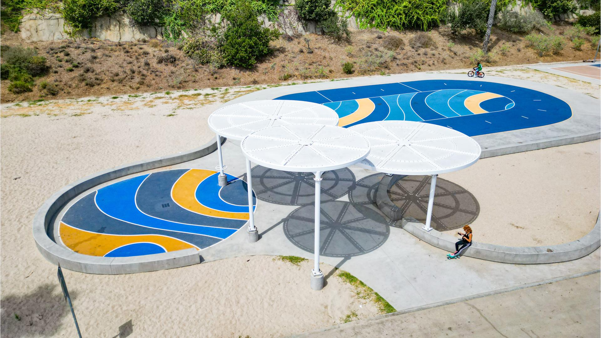 Elevated view of large circular shade structures covering a sidewalk surrounded by sand and a two additional circular safety surfaced covered areas for biking and rollerblading.